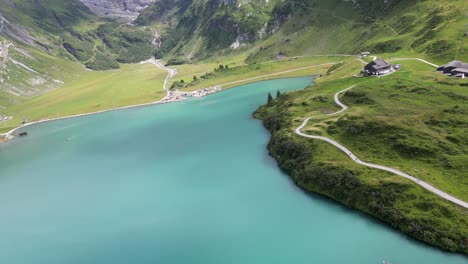 FPV-view-of-an-alpine-clear-water-lake-in-the-swiss-alps,-sinuous-stone-path-for-bikers-and-families,-mountains-behind,-Obwalden,-Engelberg