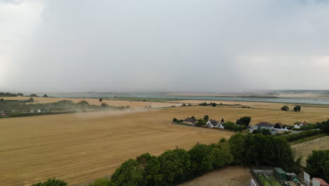 Aerial-footage-of-dust-coming-from-farmer-working-on-the-fields-in-the-summer,-harvesting-wheat-fields-in-the-UK