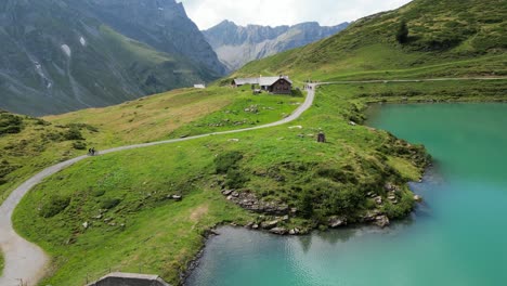 Drone-view-of-the-shore-of-an-alpine-calm-water-lake,-biking-path-and-rocky-mountains,-Engelberg,-Obwalden