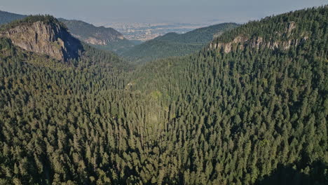 Mexico-City-Aerial-V98-Elevation-Shot-Drohne,-Die-Eine-Wunderschöne-Berglandschaft-Einfängt,-Die-Mit-Immergrünen-Nadelwäldern-Bedeckt-Ist,-Mit-Stadtbild-Im-Fernen-Hintergrund---Aufgenommen-Mit-Mavic-3-Cine---Januar-2022