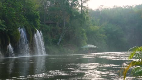Ein-Wunderschöner-Blick-Auf-Schwimmende-Häuser-Auf-Einem-Fluss-Und-Einen-Atemberaubenden-Wasserfall-Mitten-Im-Dschungel-Des-Sai-Yok-Nationalparks-In-Thailand-Mit-Dem-Hübschen-Morgennebel-Und-Der-Exotischen-Stimmung