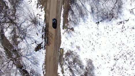 Top-Down-Aerial-Drone-Footage-of-a-Black-Sedan-Driving-Down-a-Dirt-Road-in-a-Snow-Covered-Forest
