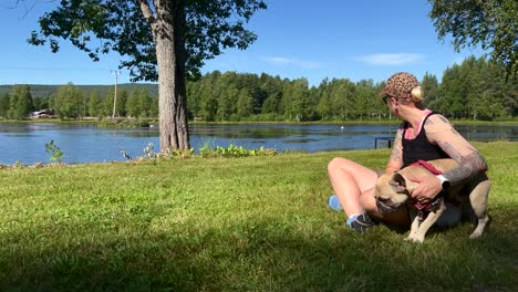A-woman-sitting-in-the-green-grass-petting-her-French-Bulldog-in-front-of-a-lake-on-a-sunny-summer-day