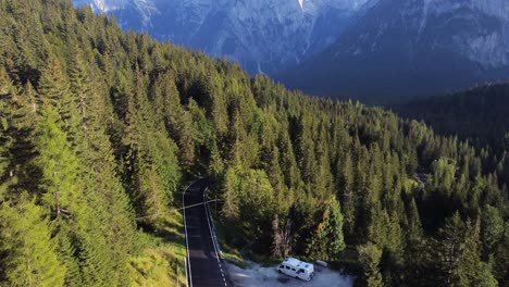 A-drone-shot-of-a-charming-mountain-road-in-the-Dolomites,-with-an-auto-camper-parked-at-the-side-of-the-road-in-the-middle-of-a-pine-tree-forest-surrounded-by-mountains-in-north-Italy