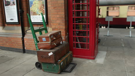 Antike-Vintage-Gepäckkoffer-Auf-Einem-Trolly-Auf-Dem-Bahnsteig-An-Einem-Bahnhof-In-England-Mit-Einer-Roten-Telefonzelle