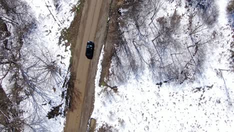Top-Down-Aerial-Drone-Footage-of-a-Black-Car-Driving-Down-a-Dirt-Road-in-the-Snowy-Wilderness