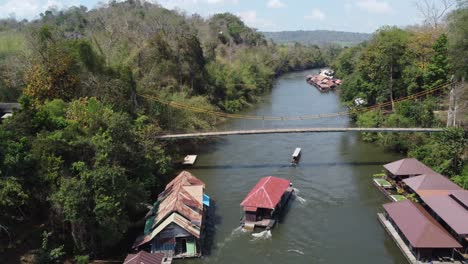 Eine-4k-drohnenaufnahme-Eines-Schwimmenden-Hauses,-Das-Auf-Einem-Fluss-Im-Sai-Yok-Nationalpark-Segelt,-An-Einem-Charmanten-Schwimmenden-Dorf-Mitten-Im-Dschungel-Vorbeifährt-Und-Unter-Einer-Brücke-In-Thailand-In-Se-Asien-Segelt