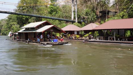 A-very-charming-wooden-floating-house-is-sailing-under-a-bridge-and-passing-a-small-floating-village-in-the-middle-of-the-jungle-of-Sai-Yok-National-Park-in-Thailand-in-Asia-on-a-clear-blue-day