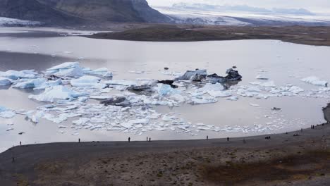 Tourist-people-standing-on-glacial-lagoon-shore-looking-at-calved-icebergs,-aerial