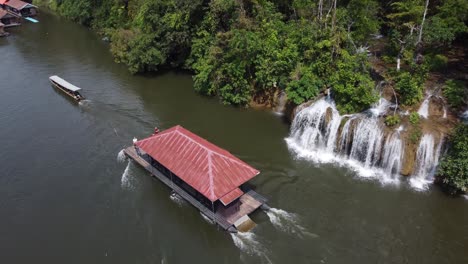 A-4K-drone-shot-of-a-floating-house-sailing-past-a-small-waterfall-in-the-jungle-of-Sai-Yok-National-Park-in-Thailand-in-Southeast-Asia