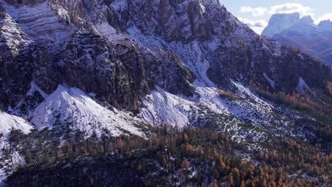 Scenic-mountain-cliff-wall-covered-in-snow-with-pine-forest-below,-aerial