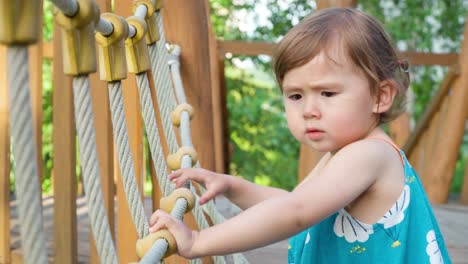 Adorable-Kid-Girl-Crossing-Wooden-Bridge-Holding-Ropes-with-Both-Hands-at-Outdoor-Playground---slow-motion-side-view