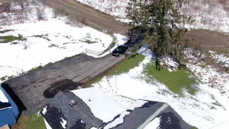 Aerial-Drone-Footage-of-a-Black-Car-Backing-Down-the-Driveway-of-an-Old,-White-House-Surrounded-by-Snow