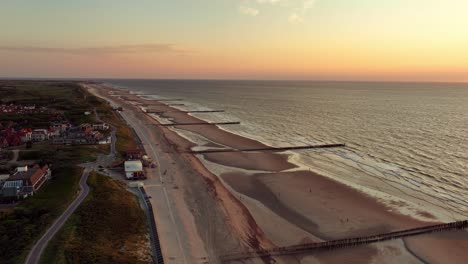Aerial-shot-of-a-small-coastal-town-along-a-beach-with-groynes-during-a-beautiful,-cloudless-sunset-in-summer