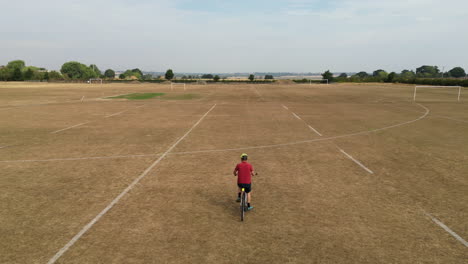Man-with-red-shirt-cycling-across-a-playing-field-in-the-UK,-drought-conditions