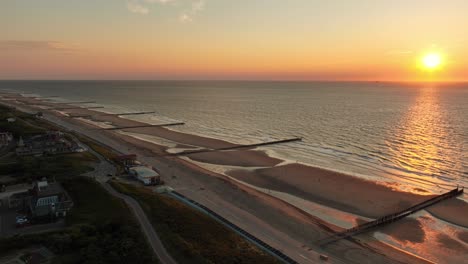 Aerial-shot-flying-over-a-small-coastal-town-along-a-beach-with-groynes-during-a-beautiful,-cloudless-sunset-in-summer