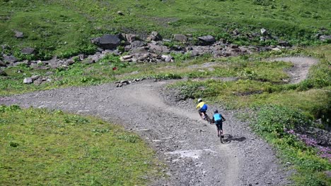 Two-riders-are-going-downhill-with-their-bikes-on-a-dirt-and-gravel-path-next-to-meadows-in-the-swiss-alps,-Obwalden,-drone-view