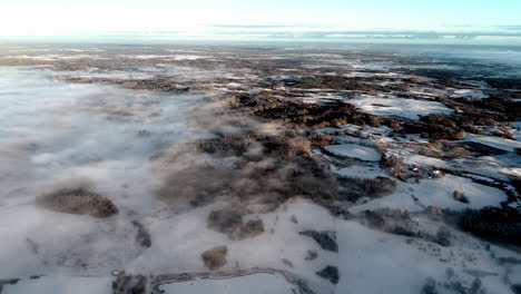 Aerial-drone-flying-through-clouds-over-snow-covered-landscape-with-coniferous-trees-during-sunrise