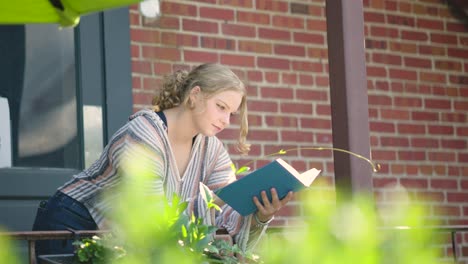 Girl-reading-book-outside-leaning-over-a-side-rail-against-a-brick-building