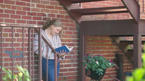 Girl-student-smiles-while-reading-a-book-outside-against-a-brick-wall