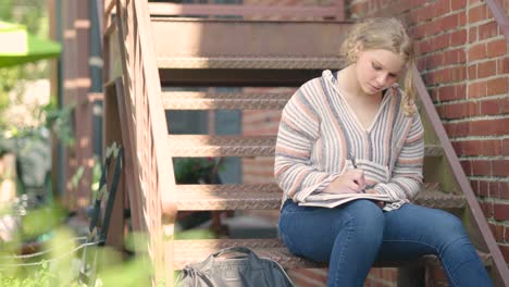 Girl-student-writes-on-notepad-sitting-on-stairs-outside