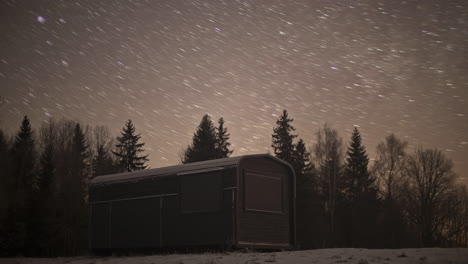 Timelapse-shot-of-star-movement-over-a-small-cabin-surrounded-by-snow-covered-landscape-at-night-time