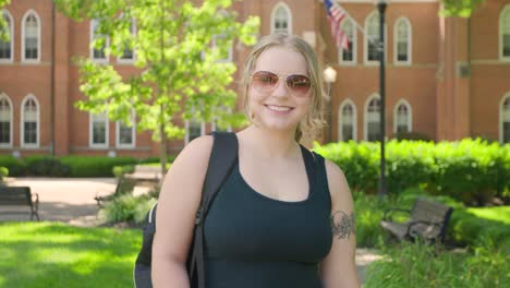 Girl-student-with-a-backpack-wearing-sunglasses-smiles-in-front-of-a-university