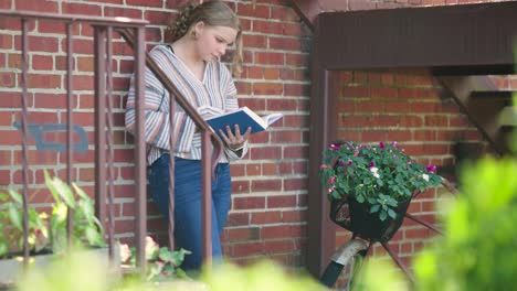Mädchen-Liest-Buch,-Während-Sie-Sich-Neben-Einem-Mit-Blumen-Geschmückten-Fahrrad-An-Eine-Mauer-Und-Eine-Treppe-Lehnt