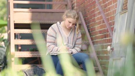 Girl-student-writes-on-notepad-while-sitting-on-metal-stairs-outside