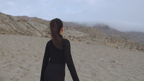 Tracking-shot-of-a-beautiful-and-mysterious-woman-from-behind-dressed-in-black-walking-through-the-desert-while-the-wind-blows