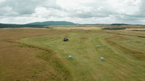 Field-full-of-round-hay-bales-with-tractor-machine-for-haybale-making,-aerial