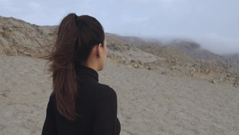 Close-up-tracking-of-a-beautiful-woman-from-behind-dressed-in-black-walking-through-a-mysterious-desert-while-the-wind-blows-in-the-afternoon