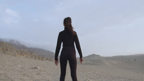 Woman-from-behind-dressed-in-black-with-a-strong-pose-standing-in-the-middle-of-the-desert-with-the-cloudy-sky-and-the-wind-blowing