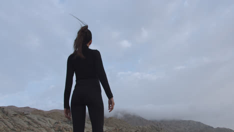 Close-up-of-a-woman-in-black-standing-strong-in-a-mysterious-desert-at-sunrise-while-the-wind-blows-and-moves-her-hair