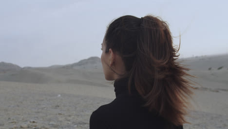 Close-up-of-the-back-of-a-woman-dressed-in-black-walking-through-the-desert-in-the-morning-while-the-wind-blows-and-moves-her-hair