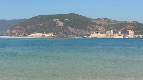 Weitwinkelaufnahme-Einer-Küstenstadt-Auf-Klippen-Mit-Grünem-Wasser,-Das-Sand-Darunter-Wäscht,-Und-Menschen,-Die-In-Booten-Segeln,-Portugal