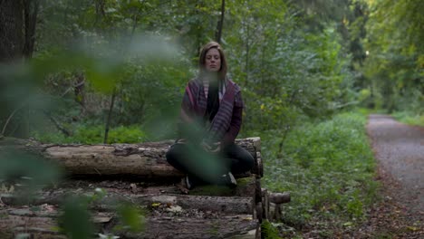 Thoughtful-natural-women-meditating-in-the-green-forest-while-sitting-on-logged-trees