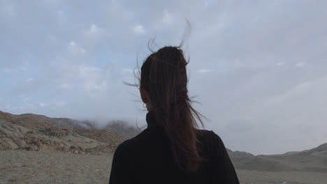 Close-up-of-the-back-of-a-mysterious-woman-dressed-in-black-standing-looking-at-the-desert-with-a-cloudy-sky-in-the-afternoon