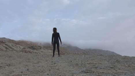 Long-shot-of-a-mysterious-woman-in-black-standing-in-the-desert-with-the-cloudy-sky-while-the-wind-blows-at-sunrise