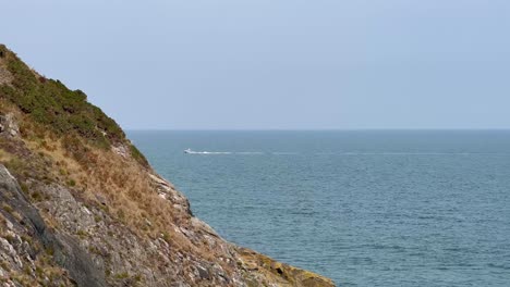 From-the-Bray-Head-cliff,-a-yacht-passes-by-in-the-distance-on-the-clam-blue-sea,-Ireland