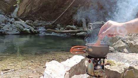Guy-cook-in-nature,-stiring-soup-on-hobo-camping-cooker-with-rivers-and-waterfall-in-background