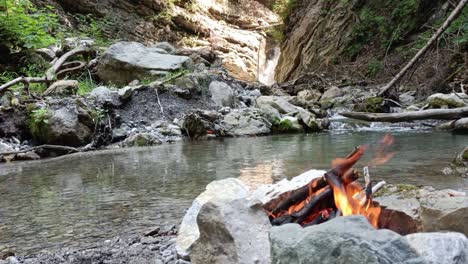 Bonfire-is-burning-peacefully-in-nature-with-no-people-around,-surrounded-by-river-and-waterfall