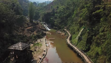 Eine-Luftaufnahme-Eines-Kleinen-Dorfes-Mit-Einer-Holzbrücke-Und-Einem-Gehweg-Entlang-Eines-Schlammigen-Braunen-Flusses-In-Sapa-Vietnam