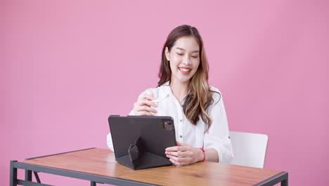 Happy-exuberant-Asian-woman-in-white-clothing-sitting-on-a-white-chair-talking-slowly-while-using-an-electronic-tablet-against-an-isolated-pink-background-2