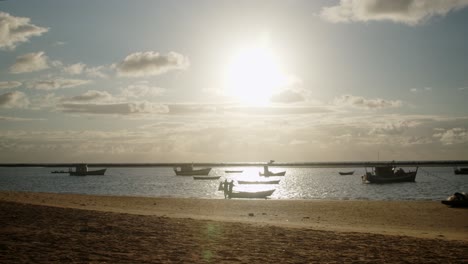 Silhouette-of-two-men-preparing-a-small-rowing-boat-to-go-fishing-and-a-woman-walking-in-the-sand-at-a-small-bay-full-of-anchored-boats-in-the-golden-hour
