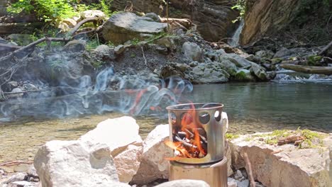 Hobo-cooker-is-burning-in-nature-with-waterfall-and-river-in-background