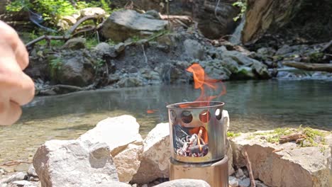 Guy-adds-wood-to-hobo-cooker-in-nature-to-cook-food,-beautiful-river-and-waterfall-in-background