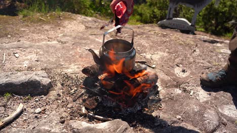 Placing-kettle-on-top-of-outdoor-bonfire-in-nature-and-adding-coffee-and-lid-before-moving-away---Sunny-day-static-hiking-clip