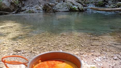 Closeup-from-hobo-cooker-cooking-tomato-soup-in-a-chinese-camping-cooker-pot-with-waterfall-and-river-in-background