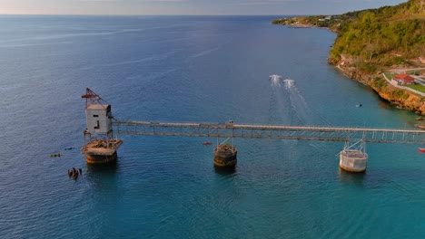 Birds-eye-view-Zoom-In-Drone-Shot-of-a-Sugar-Cane-Pier-with-jet-ski-cruising-on-the-ocean-located-in-Aguadilla,-Puerto-Rico